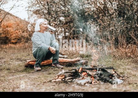 Viaggiatore donna campeggio in una foresta e rilassante vicino falò tenere tè caldo o caffè nei thermos dopo una giornata molto dura. Concetto di trekking, adv Foto Stock
