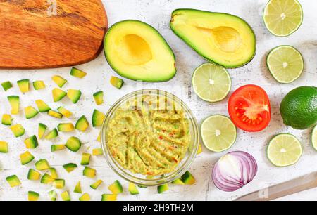 Metà e pezzi di avocado, lime di pomodoro, cipolla - guacamole base ingredienti su tavola bianca di lavoro, finito mix in piccolo recipiente di vetro vicino Foto Stock