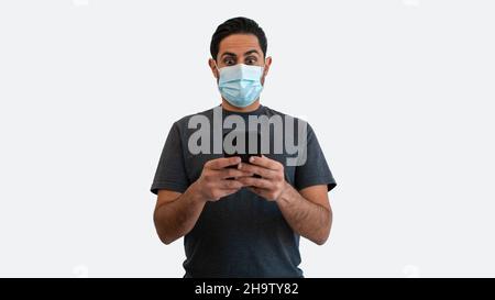 L'uomo sorpresa con l'espressione facciale scioccata sta usando lo smartphone e la maschera facciale sicura dal coronavirus su sfondo bianco. Persona eccitata che tiene un phon Foto Stock