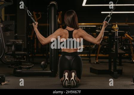 Una foto da dietro della donna sportiva che sta facendo la spinta verticale nel crossover della cabinatrice di entrambi i blocchi sulle ginocchia. Una ragazza muscolare si indossa Foto Stock