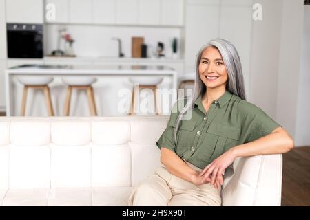 Ritratto di attraente donna di mezza età con lunghi capelli d'argento seduta in appartamento moderno, metà di età multirazziale asiatico donna riposo a casa, guardando la macchina fotografica e sorridendo con ampio sorriso toothy Foto Stock