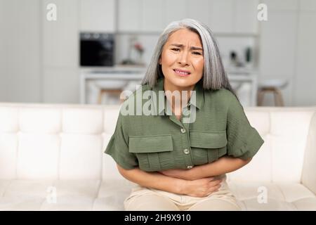 Preoccupata donna matura 50s con capelli grigi che soffrono di mal di stomaco seduta sul divano, donna di mezza età che tiene il pancino, che subisce dolore e disagio del ventre, soffrono di mestruazioni Foto Stock