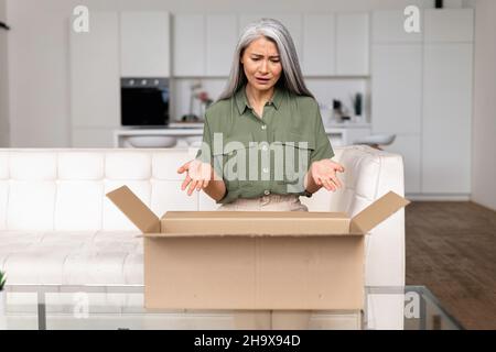 Deluso signora asiatica di mezza età sconvolto con un acquisto online. Donna dai capelli grigi che guarda all'interno della scatola di spedizione di cartone e si sente infelice e arrabbiato con ordine errato Foto Stock