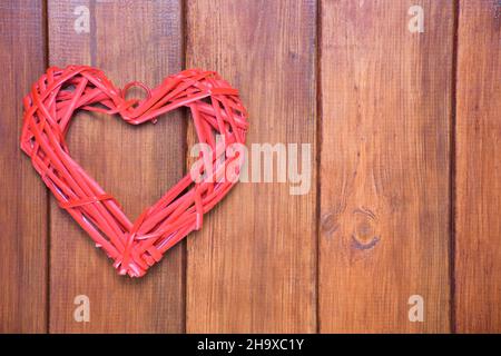 Un cuore rosso stilizzato fatto di un materiale naturale di vimini su uno sfondo di una tavola marrone di legno. Sulla destra c'è un posto per un'iscrizione. Foto Stock