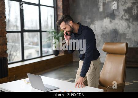 Dipendente di un ufficio di sesso maschile impegnato vicino alla scrivania dell'ufficio, che parla sullo smartphone e che controlla sul laptop, uomo d'affari in un ambiente casual e intelligente, che chiama sul cellulare, il manager ha una piacevole conversazione telefonica Foto Stock
