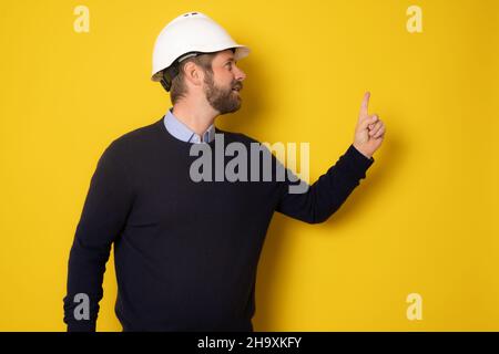 Giovane architetto uomo che indossa casco appaltatore puntando dito su isolato sfondo giallo Foto Stock