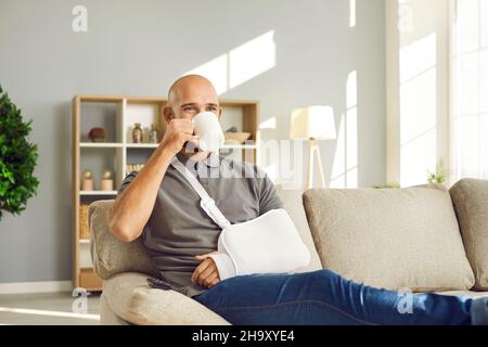 Buon giovane uomo con braccio rotto seduto sul divano a casa e una tazza di caffè Foto Stock