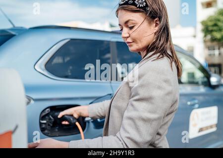 Donna che carica un'auto elettrica in ambienti urbani, Faro, Portogallo Foto Stock