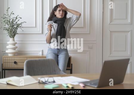 Preoccupata signora freelancer malsana controlla termometro indossando sciarpa in maglia sensazione di caldo da febbre Foto Stock