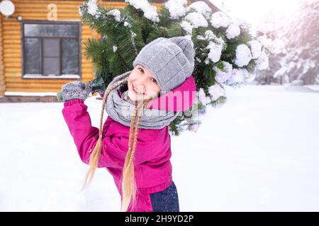 Una ragazza felice di 10-11 anni in abiti invernali porta un albero di Natale nevoso ad una casa di tronchi per decorare per la vacanza, guardando indietro, sorridendo Foto Stock
