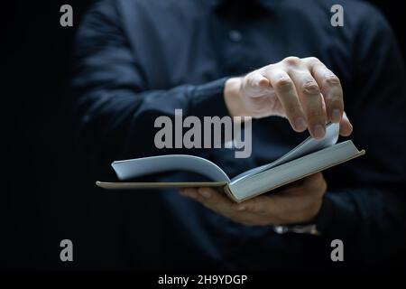 un uomo con un accappatoio nero tiene un libro davanti a lui e cerca di girare la pagina Foto Stock