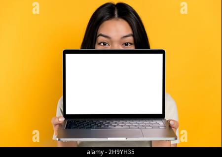 Stupito eccitato brunetta asiatica giovane donna che sbucciando fuori da dietro il laptop, guarda sorpreso la fotocamera, si erge su sfondo arancione isolato, tiene un portatile aperto con schermo bianco vuoto, spazio copia Foto Stock