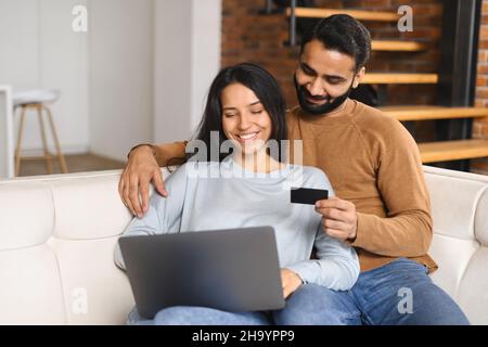 Coppia indiana allegra che guarda allo schermo del laptop e controlla il numero della carta di credito per effettuare la transazione online. Sposi sorridenti che hanno shopping in linea. Trasferimento di denaro, concetto di online banking Foto Stock