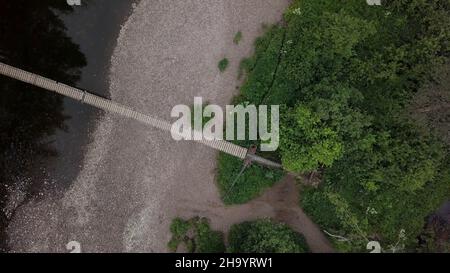 Vista dall'alto del ponte che passa attraverso il fiume nella foresta. Riprese in stock. Ponte sospeso in legno passa attraverso ampio fiume in area forestale. Foto Stock