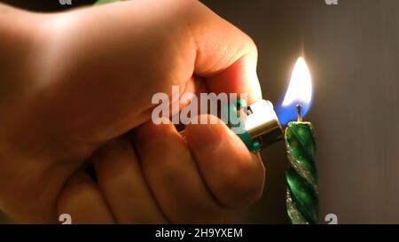 Primo piano dell'uomo che accende la candela di Natale. Concetto. L'uomo accende la candela lunga di festa per la cena intima sullo sfondo di luci sfocate. Foto Stock