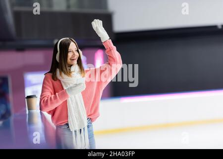 donna felice in cuffie con cuffia che tiene lo smartphone e agitare la mano sulla pista di ghiaccio Foto Stock