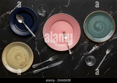 Set di piastre in porcellana con coltelli e forcelle in acciaio inox, vetri in cristallo con acqua e cucchiai sulla superficie in marmo nero Foto Stock