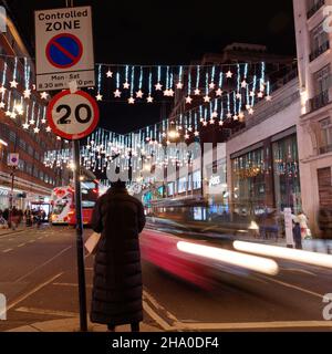 Londra, Greater London, Inghilterra, dicembre 04 2021: Lady si trova a Oxford Street mentre il traffico sibila di notte con le luci di Natale in mostra. Foto Stock