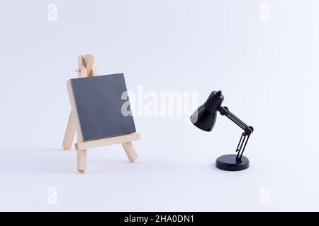 Easel in legno in miniatura con tela quadrata nera vuota per artisti e pittori - Mockup. Mini supporto in legno con tavola da disegno pulita e piccola lampada da tavolo nera su sfondo bianco, Copy Space Foto Stock