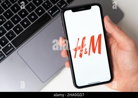 Logo dell'app H e M sullo schermo dell'iPhone per lo shopping online con un laptop sullo sfondo. Novembre 2021, San Francisco, USA Foto Stock