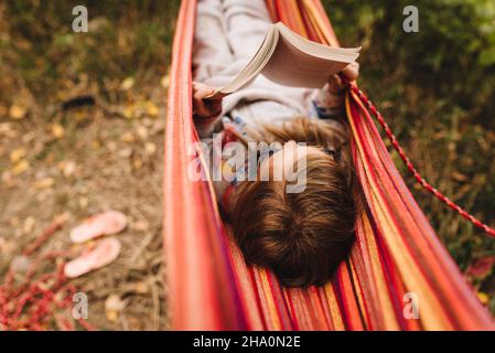 La bambina legge un libro in amaca in un caldo giorno di caduta mentre campeggio Foto Stock