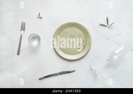 Piatto in porcellana verde con posate in acciaio inox, vaso in vetro con acqua e foglie d'oliva su tavola in marmo bianco Foto Stock