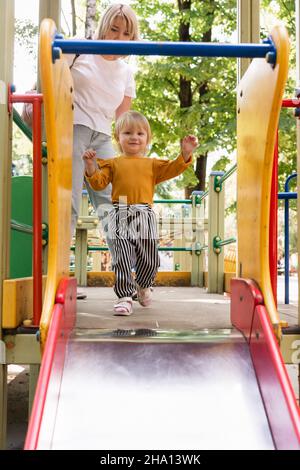 Una bella ragazza allegra cavalca uno scivolo cittadino per bambini nel parco. La mamma aiuta a non cadere. Stile di vita Foto Stock