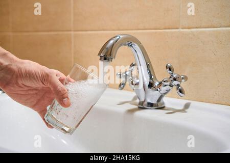 Man riempire il bicchiere d'acqua dal rubinetto a colonna in acciaio inox. La mano del maschio versa l'acqua in vetro dal rubinetto del cromo del bagno per bere l'acqua corrente Foto Stock