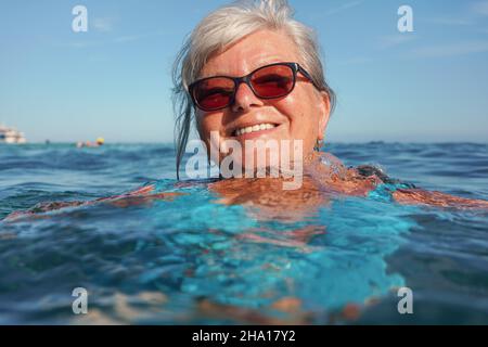 Anziana donna anziana con capelli grigi, nuoto in mare in giorno di sole, vicino al suo viso sorridente Foto Stock