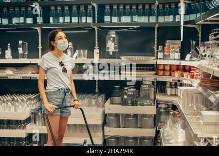 14 giugno 2021, Ufa, Russia: Donna in maschera protettiva durante la pandemia covid sceglie e acquista un vetro e altri utensili da cucina nella cenere IKEA Foto Stock