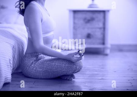 Sorridente giovane donna marrone in pigiama che pratica yoga in luminosa camera da letto al mattino. Colore di 2022 anni, molto peri, tonico Foto Stock