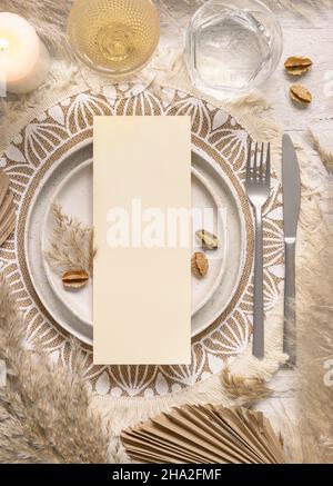 Posto da tavolo per matrimoni con carta bianca verticale sul piatto con vista dall'alto sul placemat bohémien. Boho menu di nozze carta mockup. Sagoma beige tropicale con asciugato Foto Stock