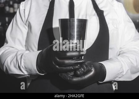 Le mani di un barista professionista in guanti di lattice nero contengono uno strumento per mescolare e preparare cocktail shaker. Foto Stock