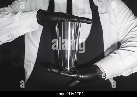 Le mani di un barman professionale in guanti di gomma di lattice contengono uno strumento per preparare e mescolare cocktail shaker alcolici, primo piano. Foto Stock