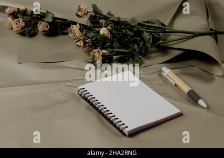 XzPrimavera notebook, penna e fiori sbiaditi. Un bouquet morente di rose gialle sullo sfondo. Foto colorata. Messa a fuoco selettiva. Foto Stock