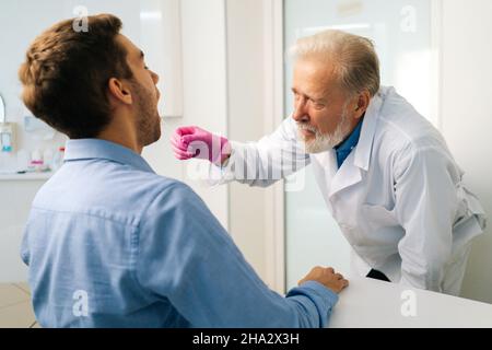 Primo piano vista torna a adulto maturo maschio medico test malato giovane paziente per possibile infezione da coronavirus utilizzando tampone di gola in ufficio medico. Foto Stock