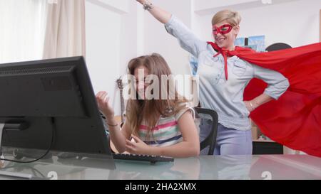 Madre in costume di supereroe viene ad aiutare la sua figlia sconvolta mentre ha problema di lavoro. Genitore gioioso con potere indossare abiti eroe motivante bambino. Concetto di genitorialità Foto Stock