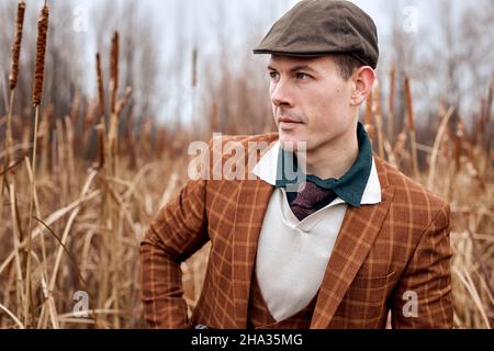 cacciatore con fucile in piedi in natura guarda fuori per la partita, nella stagione autunnale. giovane maschio caucasico in abito marrone alla moda, in campagna, in attesa, preparazione Foto Stock