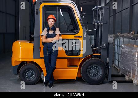 donna sorridente lavoratore di lavoro vicino a carrello elevatore a forche in fabbrica industria logistica magazzino di spedizione, in posa con le braccia ripiegate, vestito in uniforme e orang Foto Stock