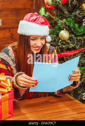 Bella Donna bianca in un casual caldo pullover e Santa Hat lettura di una scheda di Natale accanto all'albero di Natale con diverse sfere di glitter . La giovane Signora ama leggere i saluti di Natale. Coperchio vuoto. Spazio copia. Primo piano. Legno sfondo. Foto di alta qualità Foto Stock