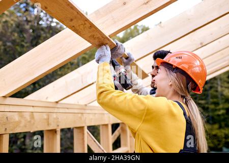 Due giovani appaltatori che lavorano con il trapano avvitatore. Carpentiere che lavora su legno Casa Skeleton telaio sezione del tetto. Tema dell'industria delle costruzioni. Femmina poll Foto Stock