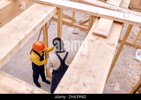 Vista dall'alto su due architetti ingegneri costruttori che lavorano insieme, costruzione casa cottage in legno. I carpentieri giovani che lavorano duro in abiti speciali lavorano un Foto Stock