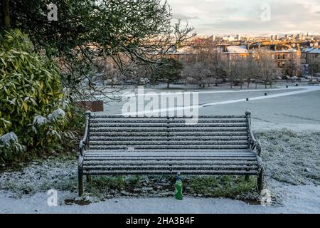 Una panca parco non occupata e una bottiglia di birra vuota al suolo coperta di neve in una giornata invernale a Queens Park, Glasgow. Foto Stock