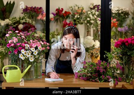 Donna sorridente Florist piccolo negozio di fiori di piccola impresa Proprietario parlare al telefono, prendere gli ordini per il negozio. Gardener femmina che rileva l'ordine del cliente durante il telefono cellulare C. Foto Stock