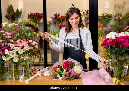 Fiorista al lavoro: Bella donna caucasica che arrangiano decorazioni floreali, facendo bouquet per i clienti. Adorabile signora in grembiule è in piedi dietro lavoro scrivania Foto Stock