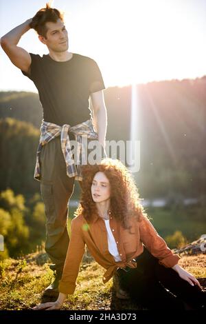 coppia caucasica sognante viaggia intorno alle montagne. Relax, riposo in piedi insieme su collina in campagna, posa al sole, vestito casual Foto Stock