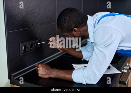 Black american Electrician ripara le prese dell'impianto elettrico residenziale, usando l'attrezzo del cacciavite. Esperto di fiducia africano Handyman in blu ouna Foto Stock