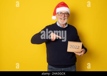 Uomo sorridente anziano che indossa il cappello di santa mostrando il calendario isolato su sfondo giallo. Foto Stock