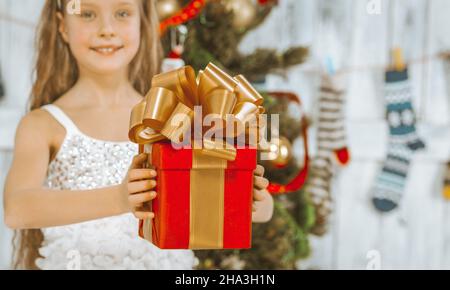 Bella ragazza di 8 anni in un abito bianco principessa contiene una scatola regalo rossa con un nastro d'oro e un grande aratro a nastro su di esso. Stocking di Natale, albero di Natale, decorazioni di Natale. Primo piano. Foto di alta qualità Foto Stock