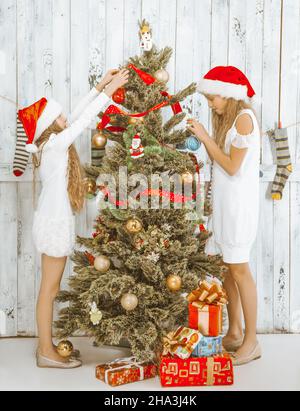 Bambini di Natale. Due ragazze teen in cappelli di Santa e vestiti bianchi stanno decorando un albero di Natale. Ci sono molti regali di diverse dimensioni e forme sotto l'albero in una grande stanza della luce. Legno sfondo. Lunghezza completa. Foto di alta qualità Foto Stock